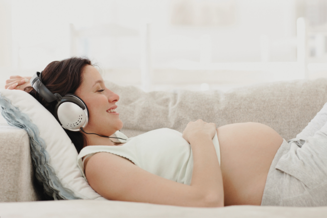 Schwangere Frau liegt auf der Couch und lächelt, weil sie eine Hypnose aus dem Hypnobirthing und Selbstliebe Audiokurs hört